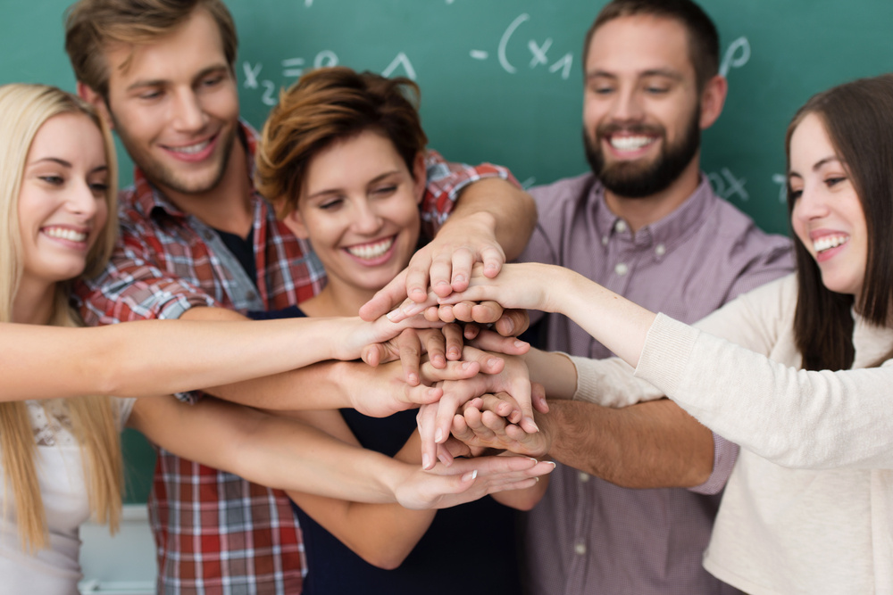 Teamwork and collaboration amongst students with a laughing group of diverse young men and women standing close together forming a stack with their hands